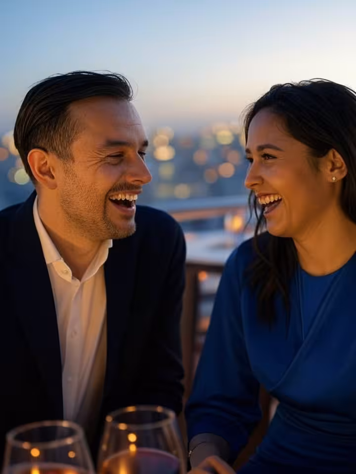 Sophisticated couple laughing genuinely during conversation at exclusive rooftop bar, city lights ba